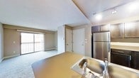 This single-floor space showcases an open-concept design that seamlessly integrates the kitchen and living area into one cohesive environment. The kitchen is equipped with a stainless steel refrigerator, dark espresso-toned cabinetry overhead, and a countertop that extends generously to provide ample workspace and casual dining potential. A stainless steel sink with a chrome faucet is positioned within the counter, completing the functional kitchen setup. Recessed lighting illuminates the kitchen area, ensuring a bright and practical workspace.

The living area transitions naturally from the kitchen, featuring neutral wall-to-wall carpeting that adds warmth underfoot. Vaulted-style ceilings enhance the sense of vertical space throughout. A large sliding glass door dominates one wall of the living area, flooding the space with natural light and providing direct access to a private balcony with outdoor views. The entryway is defined by a solid interior door alongside a closet, offering convenient storage. Neutral tones throughout the walls and flooring create a clean, adaptable aesthetic suitable for a variety of furnishing styles.