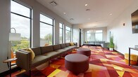 Located at 12201 Larchmere Blvd in Cleveland's Larchmere neighborhood, this single-floor community space is designed with a modern, energetic aesthetic that balances comfort and functionality. The lounge area is the centerpiece of the space, distinguished by a striking geometric carpet in vibrant shades of red, orange, and yellow that anchors the room with bold visual energy. Large floor-to-ceiling windows line the walls, flooding the interior with natural light and offering expansive views of the surrounding neighborhood and treetops. Multiple seating arrangements — including long sectional sofas, lounge chairs, and cylindrical ottoman-style seats — provide flexible areas for gathering, relaxing, or socializing. A statement chandelier and pendant lighting complement the bright, airy atmosphere. Adjacent to the lounge is a dining area with a butcher-block communal table and sleek black seating, positioned near glass double doors that open directly onto an outdoor terrace, seamlessly blending indoor and outdoor living. The kitchen area features stainless steel appliances, subway tile backsplash, granite countertops, and ample cabinetry, making it well-suited for entertaining. Curated artwork adds personality throughout, completing this inviting and thoughtfully designed communal space.
