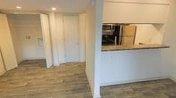 This single-floor space offers a clean, functional layout designed for comfortable everyday living. The interior is finished with warm wood-style plank flooring that runs continuously throughout, lending a cohesive and polished look to the entire unit. Neutral white walls and recessed lighting create a bright, airy ambiance that enhances the sense of openness.

The kitchen is positioned alongside a half-wall partition, providing a semi-open connection to the adjacent hallway and living areas. It features stainless steel appliances, including a refrigerator and over-the-range microwave, complemented by dark granite-style countertops that add a touch of sophistication. The design allows for easy interaction between the kitchen and the rest of the living space.

A wide hallway leads toward the remainder of the unit, where louvered bi-fold closet doors offer practical storage solutions. The entry door is solid and simply framed, contributing to the clean, uncluttered aesthetic of the space. White baseboards trim each room neatly, reinforcing the well-kept and move-in-ready condition of the interior. Overall, the space presents a tidy, modern atmosphere with thoughtful finishes and a straightforward, efficient floor plan.