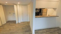 This single-floor space offers a clean, functional layout designed for comfortable everyday living. The interior is finished with warm wood-style plank flooring that runs continuously throughout, lending a cohesive and polished look to the entire unit. Neutral white walls and recessed lighting create a bright, airy ambiance that enhances the sense of openness.

The kitchen is positioned alongside a half-wall partition, providing a semi-open connection to the adjacent hallway and living areas. It features stainless steel appliances, including a refrigerator and over-the-range microwave, complemented by dark granite-style countertops that add a touch of sophistication. The design allows for easy interaction between the kitchen and the rest of the living space.

A wide hallway leads toward the remainder of the unit, where louvered bi-fold closet doors offer practical storage solutions. The entry door is solid and simply framed, contributing to the clean, uncluttered aesthetic of the space. White baseboards trim each room neatly, reinforcing the well-kept and move-in-ready condition of the interior. Overall, the space presents a tidy, modern atmosphere with thoughtful finishes and a straightforward, efficient floor plan.