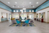 Located at 2037 Polo Gardens Dr in Wellington, Florida, this single-story community clubhouse offers a generously sized, thoughtfully designed gathering space. The heart of the facility is a grand open lounge area distinguished by soaring ceilings with a signature tray detail painted in a bold teal, adding visual depth and a resort-like ambiance throughout.

Large floor-to-ceiling glass doors and expansive windows flood the interior with abundant natural light, offering views of the outdoor pool area and lush surrounding greenery. Light-toned large-format tile flooring runs seamlessly throughout, enhancing the sense of openness. The main lounge is furnished with plush gray sofas, vibrant teal accent chairs, and a vivid geometric area rug, creating an inviting, modern atmosphere.

A dining nook features a warm wood table with yellow upholstered chairs, while a sleek bar-height counter with modern stools serves as a casual workspace or social hub. Adjacent to the lounge, a mirrored fitness center is visible, adding to the multi-functional nature of the space.

The entry foyer features a bold yellow accent niche with decorative display shelving and a mirror, setting an energetic tone from the moment of arrival. Glass-paneled private office spaces toward the rear provide a quieter setting for business or administrative use. Tall potted plants placed throughout contribute a fresh, natural element to the overall design.