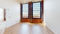 Located at 255 Promenade St in Providence, Rhode Island, this one-bedroom, one-bathroom loft-style unit showcases a compelling blend of historic industrial character and modern finishes. The open-plan living, dining, and kitchen area is the heart of the space, distinguished by soaring ceilings with arched detailing, warm hardwood flooring, and dramatic floor-to-ceiling multi-pane windows framed by exposed red brick columns. These oversized windows flood the interior with abundant natural light and offer sweeping views of the Providence skyline. The kitchen is thoughtfully designed with light wood cabinetry, open shelving, stainless steel appliances including a refrigerator and dishwasher, a sleek backsplash, and track lighting overhead. The spacious primary bedroom continues the industrial aesthetic with exposed brick accents, large industrial-style windows, and generous closet space provided by two dedicated closets. The bathroom is finished with elegant marble-style tile throughout, featuring a glass-enclosed walk-in shower for a clean, contemporary feel. Throughout the unit, the juxtaposition of raw brick and polished modern elements creates a distinctive urban ambiance that is both functional and visually striking.
