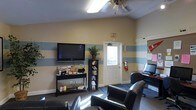 This single-floor space serves as a comfortable and functional common area designed to accommodate both leisure and light productivity. The lounge is defined by its cheerful, striped wall treatment — alternating bands of soft yellow and pale blue — that lend the space a casual, approachable character. Vaulted ceilings add a sense of openness, complemented by a ceiling fan that keeps the area comfortable year-round.

A wall-mounted flat-screen television anchors the entertainment corner, flanked by a media console and a compact shelving unit stocked with books, DVDs, and reading materials. Comfortable black leather chairs are arranged to face the screen, creating a natural gathering area for residents or visitors. Natural light filters in through a frosted-panel door, brightening the room without sacrificing privacy.

On the opposite side of the space, a computer workstation provides a functional setup for those needing access to digital resources. A bulletin board nearby offers a practical space for notices and community information. Safety features, including a fire extinguisher and illuminated exit signage, are clearly visible and accessible. The tile flooring and area rug ground the space with a clean, low-maintenance finish. Overall, the room balances utility with comfort in a well-organized, community-oriented layout.