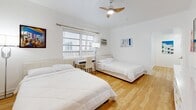 Located at 551 Michigan Ave in the heart of Miami Beach, this single-floor unit offers a clean, bright, and thoughtfully designed living space. The interior is defined by warm honey-toned hardwood flooring that runs throughout, complemented by crisp white walls that maximize the natural light pouring in through multiple windows.

The sleeping quarters are generously sized, with multiple bedrooms each accommodating two beds, a ceiling fan, a wall-mounted air conditioning unit, and a dedicated workspace. The rooms benefit from natural light through slatted blinds, with some rooms offering views of surrounding greenery.

The two bathrooms are finished in elegant marble-look tile, lending a refined, spa-like quality to the space. One bathroom features a glass-enclosed walk-in shower with mosaic tile flooring and gold-accented hardware, while the second includes a full soaking tub with a glass sliding door and a mirrored vanity unit.

The kitchen is compact yet functional, outfitted with white subway tile walls, marble-look flooring, white cabinetry, a full-size refrigerator, and a range with oven. A window above the sink provides natural ventilation. The layout also includes a dining area, rounding out this well-appointed Miami Beach residence.