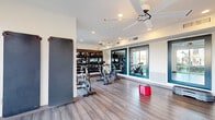 Located at 7200 N 91st Ave in Glendale, Arizona, this exercise room is a well-appointed fitness space designed to support a variety of workout routines. The room features light hardwood-style flooring throughout, lending a clean and modern aesthetic to the space.

A prominent multi-tier equipment rack lines one wall, stocked with medicine balls, TRX suspension straps, and foam rollers, providing users with a comprehensive range of functional training tools. Several stationary spin bikes are arranged across the floor, catering to cardio-focused workouts. A full-length yoga mat mounted on the wall offers additional options for stretching and floor exercises.

Large floor-to-ceiling windows along one wall flood the room with natural light and offer views of the surrounding outdoor community spaces, including green lawns and adjacent buildings. A wall-mounted display panel provides interactive fitness content, further enhancing the workout experience.

Two ceiling fans with integrated lighting ensure consistent airflow and comfort throughout the room. Teal-framed mirror panels on one wall visually expand the space and allow users to monitor their form during exercise. Recessed lighting supplements the natural light, keeping the environment bright and energizing. The room's open layout and thoughtfully selected equipment make it a functional and inviting fitness destination.