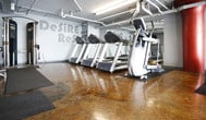 This fitness facility offers a focused and energizing environment designed for effective workouts. The space features a polished, stained concrete floor in warm amber tones that gives the interior a distinct industrial character, complemented by exposed overhead piping and ductwork that add to the raw, utilitarian aesthetic.

The cardio area is outfitted with a strong lineup of modern exercise equipment, including elliptical trainers, stair climbers, and treadmills, all arranged with ample spacing to allow comfortable use. Equipment is positioned on protective rubber mats, reflecting attention to both safety and floor preservation.

A bold red structural column serves as a visual anchor in the space, adding a striking contrast against the grey walls. Large wall-mounted mirrors extend the sense of depth throughout the room, while motivational typography — featuring phrases such as "Desire" and "Refu..." — is prominently painted across the walls, reinforcing the facility's commitment to an inspiring atmosphere.

Overhead fluorescent lighting ensures the space is well-lit throughout. The single-floor layout promotes straightforward navigation, making the facility accessible and easy to use. Overall, this gym delivers a practical, no-frills environment with quality equipment and a motivating industrial design aesthetic suited for dedicated fitness enthusiasts.