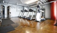 This fitness facility offers a focused and energizing environment designed for effective workouts. The space features a polished, stained concrete floor in warm amber tones that gives the interior a distinct industrial character, complemented by exposed overhead piping and ductwork that add to the raw, utilitarian aesthetic.

The cardio area is outfitted with a strong lineup of modern exercise equipment, including elliptical trainers, stair climbers, and treadmills, all arranged with ample spacing to allow comfortable use. Equipment is positioned on protective rubber mats, reflecting attention to both safety and floor preservation.

A bold red structural column serves as a visual anchor in the space, adding a striking contrast against the grey walls. Large wall-mounted mirrors extend the sense of depth throughout the room, while motivational typography — featuring phrases such as "Desire" and "Refu..." — is prominently painted across the walls, reinforcing the facility's commitment to an inspiring atmosphere.

Overhead fluorescent lighting ensures the space is well-lit throughout. The single-floor layout promotes straightforward navigation, making the facility accessible and easy to use. Overall, this gym delivers a practical, no-frills environment with quality equipment and a motivating industrial design aesthetic suited for dedicated fitness enthusiasts.