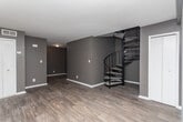 This two-floor space is defined by a consistent design palette of deep taupe and charcoal gray walls paired with white baseboards throughout. The main level features wood-look flooring in a rich dark finish, lending a grounded, modern feel to the living areas. One room on this floor showcases a wide accent wall with a subtle horizontal trim detail near the ceiling, adding a layer of architectural interest.

The kitchen is equipped with white cabinetry, light-toned countertops, a built-in sink, and white appliances including a dishwasher and refrigerator. Light wood-look flooring in the kitchen provides a bright contrast to the darker tones found elsewhere on the main level.

The upper floor is carpeted in a neutral beige tone and features multiple rooms with the same dark wall palette. A black metal stair railing connects the two levels, serving as a functional design element. Windows with horizontal blinds allow natural light to filter into the upper-level rooms, brightening the otherwise moody interior.

Overall, the space presents a cohesive, understated aesthetic with dark, neutral tones balanced by light ceilings and trim, creating a versatile environment suitable for a variety of uses.