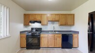 This single-floor unit offers a clean, functional layout with a neutral color palette throughout. The entry opens to a well-lit living area finished with light wood-style flooring that flows seamlessly through the main spaces, creating a bright and cohesive feel. A large window fitted with vertical blinds allows natural light to fill the room, enhancing the open atmosphere.

The kitchen is efficiently designed, featuring warm oak cabinetry extending to the upper walls for generous storage. Dark speckled countertops provide a practical work surface, complemented by a black gas range with hood ventilation, a black dishwasher, and a stainless-steel sink — a complete and functional cooking space.

The bedroom is comfortably sized, carpeted for warmth, and includes a window overlooking greenery outdoors. It connects directly to an en-suite bathroom equipped with oak vanity cabinetry, dark speckled countertops, a wide mirror, and a separate tub area. A second bathroom in the unit mirrors this design with matching vanity finishes and a tub-shower combination, offering added convenience for guests or additional occupants.