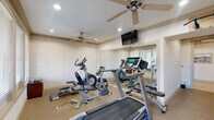 Located in Magnolia, TX, this dedicated exercise room offers a well-equipped fitness space designed for convenient, focused workouts. The room features a carpet flooring and is finished with neutral, light-toned walls accented by warm wood crown molding, creating a clean and inviting atmosphere.

The space is outfitted with multiple cardio machines, including a treadmill, elliptical trainer, and stationary bike, providing a range of aerobic exercise options. A large wall mirror spans one side of the room, visually expanding the space and allowing users to monitor their form during workouts. A wall-mounted flat-screen television adds an entertainment element to exercise sessions.

Recessed lighting and dual ceiling fans ensure the room remains well-lit and comfortably ventilated throughout use. Large windows fitted with blinds line one wall, inviting natural light while offering privacy control. A glass-panel door provides direct access to an outdoor area, adding convenience and the option for fresh air.

Overall, this exercise room delivers a practical, self-contained fitness environment with thoughtful design touches that balance functionality and comfort.