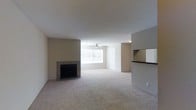 This single-floor space centers around a spacious, open-plan living area finished with wall-to-wall carpeting in a neutral tone. A tile-surround fireplace serves as a focal point along one wall, adding a functional and aesthetic element to the room. Natural light filters in through a window fitted with horizontal blinds, brightening the interior and contributing to the overall airy feel. A ceiling fan is centrally positioned, providing both ventilation and ambient lighting. The neutral beige walls and white ceiling create a clean, understated palette throughout. A pass-through window with a dark countertop connects the living area to the adjacent kitchen space, facilitating an open flow between the two areas. A doorway visible toward the rear of the living area leads to additional rooms within the unit. The overall design is practical and straightforward, with a cohesive neutral color scheme that provides a versatile backdrop for a variety of interior arrangements.