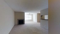 This single-floor space centers around a spacious, open-plan living area finished with wall-to-wall carpeting in a neutral tone. A tile-surround fireplace serves as a focal point along one wall, adding a functional and aesthetic element to the room. Natural light filters in through a window fitted with horizontal blinds, brightening the interior and contributing to the overall airy feel. A ceiling fan is centrally positioned, providing both ventilation and ambient lighting. The neutral beige walls and white ceiling create a clean, understated palette throughout. A pass-through window with a dark countertop connects the living area to the adjacent kitchen space, facilitating an open flow between the two areas. A doorway visible toward the rear of the living area leads to additional rooms within the unit. The overall design is practical and straightforward, with a cohesive neutral color scheme that provides a versatile backdrop for a variety of interior arrangements.