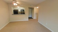 This single-floor space offers a functional and well-organized layout designed for comfortable everyday living. The open-concept living area is finished with plush wall-to-wall carpeting in a warm neutral tone, creating a welcoming atmosphere throughout. A ceiling fan with an integrated light fixture is centrally positioned in the living area, providing both air circulation and overhead lighting.

The kitchen is accessible via a raised pass-through counter that opens directly into the living space, making it ideal for both cooking and entertaining. White cabinetry lines the kitchen walls, offering ample storage, while black appliances — including a refrigerator, microwave, and range — provide a clean contrast. Dark countertops complete the kitchen's practical aesthetic.

A doorway from the main living area leads further into the unit, providing access to additional rooms. A wall-mounted utility panel, likely a thermostat or electrical access point, is visible near the hallway entrance. Neutral beige walls and white baseboards run consistently throughout, contributing to a clean, cohesive look. Ceiling vents indicate a central HVAC system for year-round climate control. Overall, the space is straightforward, efficiently arranged, and move-in ready.
