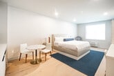 Located at 695 Merrick Ave in Westbury, NY, this thoughtfully designed studio unit offers a seamless single-floor layout that maximizes both comfort and functionality. Light oak hardwood flooring flows throughout the space, complementing a clean, contemporary color palette of soft whites and cool grays.

The open main area serves as a combined living and sleeping space, anchored by a king-sized bed with an upholstered headboard and accented by a teal area rug. A wall-mounted flat-screen TV and a sleek white dresser with geometric detailing provide practical elegance, while a marble-topped bistro table with two chairs offers a cozy sitting nook near the window, which allows natural light to fill the room.

The integrated kitchenette features flat-front white cabinetry extending to the ceiling, a stainless steel microwave, a countertop coffee maker, a compact sink with a gold-finish faucet, and a beverage refrigerator — all designed for efficient, self-sufficient living.

The bathroom is a standout feature, finished with large-format white marble-look floor tiles and gold fixtures throughout. A open vanity with a marble countertop and gold-framed mirror adds sophistication, while a glass-enclosed walk-in shower with marble tile surround completes the spa-inspired aesthetic. A louvered closet door at the entry provides additional storage, rounding out this well-appointed space.