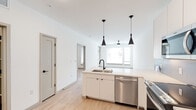 This single-floor space showcases a clean, contemporary design with an open-concept layout that seamlessly connects the kitchen and living areas. Light hardwood flooring runs throughout, lending warmth and cohesion to the bright, white-walled interior.

The kitchen is a standout feature, centered around a peninsula island fitted with a double undermount sink, matte black faucet, and pendant lighting. White flat-panel cabinetry extends to the ceiling, complemented by a subway tile backsplash and a full suite of stainless steel appliances, including a side-by-side refrigerator with ice and water dispenser, electric range, over-the-range microwave, and dishwasher.

The living area is generously proportioned, bathed in natural light from large windows fitted with blinds, and equipped with a ceiling fan. A stylish barn-style sliding door adds a modern architectural detail to the space.

The bathroom features classic white subway tile in the tub surround, a built-in open shelving unit, a white vanity, and continued hardwood-style flooring for a consistent aesthetic.

A dedicated laundry area houses a stacked washer and dryer, with an adjacent half bath and closet with hanging storage, offering practical convenience within a well-organized floor plan.