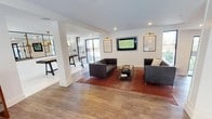 This single-floor amenity space serves as a well-appointed lounge and entertainment destination. The entrance is marked by sleek, black-framed glass doors with etched signage, offering a clear view of the adjacent outdoor pool courtyard beyond a secured gate.

Inside, the main lounge area is anchored by a pair of dark tufted leather sofas arranged around a glass-top coffee table, set atop a warm-toned area rug. A wall-mounted flat-screen television is flanked by sconce lighting and framed artwork, creating a polished focal point. Recessed ceiling lighting and large windows allow natural light to fill the space, complementing the light wood-tone flooring that runs throughout.

Adjacent to the lounge, a partially visible game room features what appears to be a billiards or shuffleboard table, separated by a white structural column that defines the two zones while maintaining an open, connected feel. Pendant lighting in the game area adds a distinct ambiance. The overall design aesthetic is contemporary and clean, with a neutral white palette offset by warm wood tones and dark furniture accents, creating an inviting and functional communal environment.