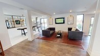 This single-floor amenity space serves as a well-appointed lounge and entertainment destination. The entrance is marked by sleek, black-framed glass doors with etched signage, offering a clear view of the adjacent outdoor pool courtyard beyond a secured gate.

Inside, the main lounge area is anchored by a pair of dark tufted leather sofas arranged around a glass-top coffee table, set atop a warm-toned area rug. A wall-mounted flat-screen television is flanked by sconce lighting and framed artwork, creating a polished focal point. Recessed ceiling lighting and large windows allow natural light to fill the space, complementing the light wood-tone flooring that runs throughout.

Adjacent to the lounge, a partially visible game room features what appears to be a billiards or shuffleboard table, separated by a white structural column that defines the two zones while maintaining an open, connected feel. Pendant lighting in the game area adds a distinct ambiance. The overall design aesthetic is contemporary and clean, with a neutral white palette offset by warm wood tones and dark furniture accents, creating an inviting and functional communal environment.