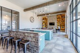 This thoughtfully designed single-floor space masterfully blends rustic and contemporary elements into a cohesive, inviting environment. Exposed wooden ceiling beams run the length of the open-plan interior, adding warmth and architectural character, while large black-framed floor-to-ceiling windows flood the space with abundant natural light and offer expansive outdoor views of the surrounding area.

The lounge area serves as a central gathering point, featuring plaid upholstered sofas, eclectic patterned armchairs, and a reclaimed wood coffee table arranged around a striking stacked-stone accent wall. Built-in open shelving, bathed in warm amber tones, frames a wall-mounted television and displays carefully curated decorative objects, enhancing the space's artistic character.

The kitchenette area is a standout feature, showcasing bold teal cabinetry paired with white countertops and brick-accented detailing. Industrial-style pendant lighting hangs above the central island, complementing the mixed-material aesthetic. Bar-height seating with industrial stools lines a counter adjacent to the kitchen, creating a casual dining and gathering area.

The geometric-patterned tile flooring ties all zones together seamlessly, while steel-framed glass partitions add a modern industrial touch. Overall, the space exudes a warm yet contemporary ambiance ideal for social interaction.