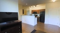 This single-floor residence offers a thoughtfully designed open-concept layout that seamlessly connects the living and kitchen areas. The kitchen is well-appointed with rich medium-toned wood cabinetry, black appliances including a full-size refrigerator, microwave, and range, complemented by a white peninsula counter that provides additional prep space and serves as a natural divider between the kitchen and living area. Track lighting overhead ensures the kitchen is well-lit and functional. The living area extends comfortably from the kitchen, featuring warm hardwood flooring that runs throughout and lends a cohesive, inviting feel to the space. A dedicated area accommodates a media console and television, making it well-suited for everyday living. A walk-in closet or storage space is visible in the background, indicating thoughtful storage solutions within the floor plan. The neutral wall tones and light-filled interior contribute to a clean, airy ambiance. Overall, this space offers a practical and comfortable layout with a balance of style and functionality.