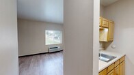 Located at 2620-2626 Chippewa St in St. Louis, Missouri, this single-floor unit offers a practical and thoughtfully designed layout. The main living area features light wood plank flooring throughout, neutral beige walls, and dark baseboards that provide a clean, modern contrast. A window with blinds allows natural light to filter in, and a wall-mounted HVAC unit ensures year-round climate control. A pass-through opening connects the living space to the kitchenette, which is equipped with wooden cabinetry, a sink, and a range, making efficient use of the available space. The hallway connects the main living area to the bathroom and includes a sliding wood door and a dedicated closet area with a clothes rod for storage. Track lighting in the hallway provides additional illumination. The bathroom is equipped with an accessible tub-shower combination featuring multiple grab bars and a handheld showerhead, underscoring a commitment to accessibility and safety. Throughout the unit, the consistent use of light tones and wood-finish flooring creates a cohesive, clean aesthetic. The overall design prioritizes functionality and accessibility within a compact, efficiently organized footprint.