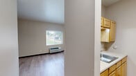 Located at 2620-2626 Chippewa St in St. Louis, Missouri, this single-floor unit offers a practical and thoughtfully designed layout. The main living area features light wood plank flooring throughout, neutral beige walls, and dark baseboards that provide a clean, modern contrast. A window with blinds allows natural light to filter in, and a wall-mounted HVAC unit ensures year-round climate control. A pass-through opening connects the living space to the kitchenette, which is equipped with wooden cabinetry, a sink, and a range, making efficient use of the available space. The hallway connects the main living area to the bathroom and includes a sliding wood door and a dedicated closet area with a clothes rod for storage. Track lighting in the hallway provides additional illumination. The bathroom is equipped with an accessible tub-shower combination featuring multiple grab bars and a handheld showerhead, underscoring a commitment to accessibility and safety. Throughout the unit, the consistent use of light tones and wood-finish flooring creates a cohesive, clean aesthetic. The overall design prioritizes functionality and accessibility within a compact, efficiently organized footprint.