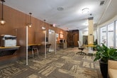 Located at 9000 Beaver Creek Rd in Alpharetta, Georgia, this single-floor amenity space offers a well-appointed and functional environment designed for both productivity and relaxation. The space is anchored by a dedicated business center equipped with multiple computer workstations set atop warm yellow-toned desks, complemented by ergonomic seating and privacy partitions — ideal for focused work or administrative tasks. A striking spiral staircase serves as an architectural focal point in the business center area, adding visual interest to the otherwise clean, contemporary layout.

Throughout the space, a distinctive patterned carpet featuring organic, branch-like motifs in neutral tones of beige, gray, and taupe creates a cohesive and sophisticated aesthetic. The walls feature rich, textured brown paneling accented by curated artwork, adding warmth and depth to the interiors.

Transitioning into the lounge area, comfortable seating arrangements include sofas and armchairs in muted, complementary tones, centered around a flat-screen television and accented by lush indoor plants. Large windows allow ample natural light to fill the space and provide views of the surrounding greenery. A more intimate seating nook features a textured sculptural wall piece and cozy individual seating, offering a quiet retreat within the broader space. The overall ambiance is polished, welcoming, and thoughtfully designed.