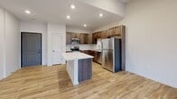 This well-designed one-bedroom, one-bathroom unit offers a thoughtful single-level layout that maximizes its efficiently used floor space. The open-concept kitchen and living area serves as the heart of the unit, featuring light-toned luxury vinyl plank flooring throughout and recessed lighting overhead. The kitchen is outfitted with rich, medium-toned wood cabinetry, quartz countertops, a subway tile backsplash, and a full suite of stainless steel appliances including a refrigerator, range, microwave, and dishwasher. A central kitchen island with a quartz top provides additional prep space and casual seating. The living area opens directly onto a private balcony through sliding glass doors, complemented by a side window that draws in natural light. The bedroom is a comfortable size, finished with plush carpet, a ceiling fan with light fixture, and a window with outdoor views. A dedicated closet provides organized storage. The bathroom features a full tub-shower combination, a wood-tone vanity with a quartz countertop, and matching LVP flooring, maintaining a cohesive and modern aesthetic throughout the unit.