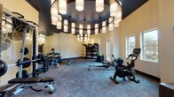 This single-level fitness center is designed with both functionality and architectural sophistication in mind. The space is defined by its striking dark ceiling, from which multiple large, tiered chandeliers hang, each fitted with cylindrical fabric shades that cast a warm, inviting glow throughout the room. The generous floor plan accommodates a comprehensive range of equipment without feeling crowded, with durable low-pile carpet flooring providing a practical yet polished surface underfoot.

The strength training area is well-stocked with a full dumbbell rack, medicine balls, TRX suspension systems, weight benches, and a squat rack loaded with plate weights. Nearby shelving units organize additional accessories and fitness tools. Full-length mirrors line one wall, visually expanding the space and assisting users during training. The cardio zone houses treadmills, a rowing machine, a stationary spin bike, and an elliptical trainer, positioned near large windows that bring in natural light and offer outdoor views.

A decorative glass partition wall featuring a bold, colorful geometric mural separates a secondary area, adding an artistic focal point. A built-in upholstered bench beneath a textured accent wall with a mounted flat-screen television provides a comfortable rest area. The overall ambiance balances upscale design with a well-equipped, purpose-driven athletic environment.
