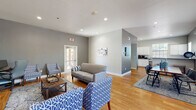 This single-floor community amenity space is thoughtfully designed to offer residents a comfortable and functional environment for both relaxation and activity. The lounge area features warm hardwood flooring, recessed lighting, and a neutral gray and white color palette. Seating is arranged around a rustic barn-door media console with a large flat-screen TV, complemented by patterned blue accent chairs, a loveseat, and a coffee table atop a patterned area rug — creating an inviting space for gathering. French doors allow natural light to flow in and provide easy access to the exterior. Adjacent to the lounge is a well-maintained kitchenette outfitted with white cabinetry, gray laminate countertops, a stainless steel sink, and a coffee station, all brightened by a window overlooking the outdoors. The fitness center is generously equipped with multiple treadmills, a cable weight machine, a stationary bike, an adjustable weight bench, a dumbbell rack, and additional cardio equipment. Durable rubber flooring, large overhead fluorescent lighting, wall-mounted mirrors, and a mounted TV complete the workout space. French doors in the fitness area provide direct outdoor access, enhancing ventilation and convenience throughout.