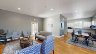 This single-floor community amenity space is thoughtfully designed to offer residents a comfortable and functional environment for both relaxation and activity. The lounge area features warm hardwood flooring, recessed lighting, and a neutral gray and white color palette. Seating is arranged around a rustic barn-door media console with a large flat-screen TV, complemented by patterned blue accent chairs, a loveseat, and a coffee table atop a patterned area rug — creating an inviting space for gathering. French doors allow natural light to flow in and provide easy access to the exterior. Adjacent to the lounge is a well-maintained kitchenette outfitted with white cabinetry, gray laminate countertops, a stainless steel sink, and a coffee station, all brightened by a window overlooking the outdoors. The fitness center is generously equipped with multiple treadmills, a cable weight machine, a stationary bike, an adjustable weight bench, a dumbbell rack, and additional cardio equipment. Durable rubber flooring, large overhead fluorescent lighting, wall-mounted mirrors, and a mounted TV complete the workout space. French doors in the fitness area provide direct outdoor access, enhancing ventilation and convenience throughout.