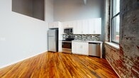 Located at 2000 E Westmoreland St in Philadelphia, PA, this single-floor unit masterfully combines industrial character with contemporary updates. Original hardwood floors run throughout, lending warmth and continuity to every room, while exposed brick walls and visible ductwork preserve the building's historic industrial identity.

The open kitchen is a standout feature, showcasing white shaker cabinetry, granite-style countertops, a mosaic tile backsplash, and a full suite of stainless steel appliances including a refrigerator, electric range, microwave, and dishwasher. Soaring ceilings and oversized arched windows in the kitchen and living area flood the space with natural light and offer open views of the surrounding Philadelphia neighborhood.

A hallway with louvered closet doors connects the living areas to the private spaces, with exposed ceiling ductwork adding to the loft-like aesthetic. The bedroom area features smooth gray walls, original hardwood flooring, and ample space for furnishings.

The bathroom is cleanly finished with large-format neutral floor tiles, a white vanity with granite countertop, and a full tub-shower combination dressed in marble-patterned subway tile. The contrast between the polished bathroom and the raw industrial elements throughout creates a compelling and cohesive aesthetic unique to this Philadelphia address.
