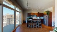 This single-floor urban residence offers a thoughtfully designed open-concept layout that seamlessly blends the kitchen and living areas. The kitchen is a standout feature, showcasing rich warm-toned cabinetry, granite countertops, a light aqua glass tile backsplash, and a full suite of stainless steel appliances including a range, microwave, dishwasher, and French door refrigerator. A rustic reclaimed wood dining table with industrial-style stools anchors the space, adding an inviting contrast to the modern finishes. Track lighting runs throughout the main living area, illuminating the warm hardwood floors below.

The living room is generously sized with elevated ceilings and a clerestory window detail that adds architectural interest. A large sectional sofa, area rug, and gallery wall create a comfortable and personalized atmosphere.

Floor-to-ceiling windows and a glass door in the main area invite abundant natural light and provide views of the surrounding urban streetscape, blurring the boundary between interior and exterior.

The bedroom is a calm, neutral retreat finished with plush carpet, soft wall tones, and a high clerestory window that allows natural light in while maintaining privacy. The overall ambiance of the space is modern yet warm, balancing contemporary urban design with cozy, livable details throughout.