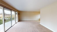 This single-floor residence offers a functional and well-organized layout designed for comfortable everyday living. The generously sized living area is finished with plush neutral carpeting and warm beige walls, creating a bright, welcoming atmosphere. A sliding glass door opens to an outdoor balcony, allowing natural light to fill the space and providing pleasant outdoor views. The kitchen is thoughtfully designed in a galley configuration, featuring rich cherry wood cabinetry, granite countertops, a stainless steel sink, and tile flooring. White appliances including a refrigerator, oven, and dishwasher complete the functional cooking space. The layout flows naturally from the kitchen into the main living area, making the home feel open and connected. Multiple bedrooms are finished with soft carpeting, ceiling fans with light fixtures, and windows fitted with blinds for privacy and light control. One bedroom includes a mirrored sliding closet door, adding depth and practicality. The primary bathroom features a frameless glass-enclosed walk-in shower with neutral tile surround and a decorative mosaic accent border, paired with a granite-topped vanity. A second full bathroom offers a tub-shower combination with frosted glass doors, tile flooring, and a matching granite vanity, providing ample convenience for residents and guests alike.