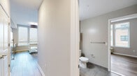 This well-appointed one-bedroom, one-bathroom unit at 26 University Place Blvd in Jersey City, NJ offers a thoughtfully designed layout with modern finishes throughout. Light wood-style flooring runs seamlessly across the entire unit, complementing the neutral gray-toned walls and crisp white trim for a clean, contemporary aesthetic.

The open-concept kitchen and living area serves as the heart of the unit. The kitchen features two-tone cabinetry — white upper cabinets paired with gray lower cabinets — accented by a white subway tile backsplash and stainless steel appliances, including a range, over-the-range microwave, and sink. Large windows flood the space with natural light and offer views of the surrounding Jersey City neighborhood.

The spacious bedroom continues the light, airy feel with a wide bank of windows and a double-door closet providing generous storage. A wall-mounted HVAC unit ensures year-round comfort.

The bathroom is finished with large-format tile flooring and includes accessibility-friendly grab bars. The unit also benefits from an in-unit laundry area and a hallway with additional closet space, rounding out a practical and comfortable living environment.