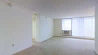 This single-floor space presents a clean and minimalist layout characterized by an open main living area with soft, neutral-toned wall-to-wall carpeting that extends throughout. The room is finished with crisp white walls and a smooth white ceiling, contributing to a bright and airy atmosphere. A large window fitted with vertical blinds dominates one wall, allowing generous amounts of natural light to fill the space. Beneath the window sits a wall-mounted air conditioning unit, complemented by a baseboard heating unit along the perimeter, ensuring year-round climate control. A partial interior wall partially divides the space, adding subtle definition without fully enclosing the layout. A doorway visible toward the rear leads to an adjoining room or corridor, suggesting additional space beyond the main living area. The overall design is straightforward and functional, with neutral finishes that provide a versatile canvas suitable for a variety of furnishing arrangements and interior styles. The absence of decorative elements keeps the focus on the spatial qualities of the unit itself.