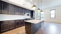 Located at 70 N High St in the heart of downtown Columbus, Ohio, this 2-bedroom, 1-bathroom residence offers a thoughtfully designed open-concept layout with modern finishes throughout. The kitchen and living area form a seamless, light-filled space anchored by a striking central island with a white quartz countertop, integrated sink, and built-in outlets. Rich, dark-stained cabinetry lines the walls, complemented by a classic white subway tile backsplash and stainless steel appliances. A trio of brass pendant lights hang above the island, adding warmth to the contemporary aesthetic. Warm hardwood-style flooring extends through the main living areas. The primary bedroom is generously sized, offering multiple large black-framed windows with views of the surrounding Columbus cityscape, and is complemented by a spacious walk-in closet outfitted with full wire shelving systems and plush carpet underfoot. The second bedroom also features hardwood-style flooring and ample natural light. The bathroom is designed with accessibility in mind, featuring grab bars, a walk-in tile shower, a navy vanity with quartz countertop, and matte black fixtures. A connecting hallway ties the layout together efficiently, creating a functional and well-organized living environment in the heart of the city.