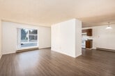 This single-floor unit offers a well-organized layout with consistent light wood-style flooring and crisp white walls throughout, creating a bright and cohesive feel. The living area is generously sized with a textured ceiling and open sightlines that extend toward a sliding glass door leading to a private balcony, where greenery is visible beyond. Natural light filters in through large windows fitted with blinds and vertical slat coverings.

The dining area is defined by a pendant light fixture overhead, with a baseboard heater and a hallway connecting the space to the rest of the unit. The kitchen features rich, dark wood cabinetry, laminate countertops, and a full suite of white appliances including a refrigerator, electric range, and dishwasher. A window above the sink brings in additional natural light.

The bedroom includes a built-in closet with shelving and is serviced by a baseboard heater, with easy access to the hallway. Two bathrooms are present — one featuring a walk-in shower with grab bars and a vanity with a dark countertop, and a second with a bathtub and vanity. Both bathrooms continue the light wood-style flooring seen throughout the unit. The overall ambiance is clean, functional, and move-in ready.
