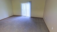 This single-floor space features an open living area finished with wall-to-wall carpeting in a neutral speckled pattern, providing a comfortable and practical foundation underfoot. The room is defined by soft yellow-toned walls and dark wood baseboards that frame the space with a clean, classic look. A prominent sliding glass door serves as a central architectural feature, fitted with sheer white curtains that diffuse natural light throughout the interior while maintaining a bright, airy atmosphere. The door opens directly onto a private balcony, visible through the glass, offering an outdoor extension of the living space. The overall layout is spacious and versatile, lending itself well to a variety of furniture arrangements. The combination of natural light from the sliding door, the neutral color palette, and the carpeted flooring creates a calm and livable environment. The space is presented in a clean, vacant condition, making it easy to assess its full dimensions and potential.