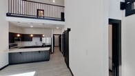 This contemporary space makes a striking first impression with its dramatic double-height ceilings and crisp white walls that amplify the sense of openness. The living area is bathed in natural light from strategically placed clerestory windows positioned high on the walls, and is accented by elegant pendant light fixtures that add warmth and character to the expansive volume.

The open-concept kitchen is a standout feature, showcasing dark espresso cabinetry paired with bright white countertops for a bold contrast. A large kitchen island provides ample workspace and casual seating, while stainless steel appliances — including a refrigerator, range, and microwave — complete the modern aesthetic. A subway tile backsplash adds texture and visual interest behind the sink and cooking area.

Light wood-tone flooring flows seamlessly throughout, complementing the space's modern palette. A bold black barn door serves as both a functional room divider and a strong design element, adding an industrial-chic touch to the interior.

The bathroom is thoughtfully designed with dark vanity cabinetry, a white countertop with an undermount sink, a framed mirror, and a tiled walk-in shower. Chrome fixtures and grab bars provide a polished, accessible finish. An upper-level mezzanine with black metal railings overlooks the main living area, adding architectural depth and visual interest to the overall design.
