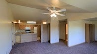 This single-floor space features an open-concept layout that seamlessly connects the living area and kitchen, creating a functional and cohesive floor plan. The living area is finished with plush, neutral-toned carpet that extends throughout, providing a comfortable underfoot feel. A ceiling fan with a built-in light fixture is centrally mounted, offering both air circulation and ambient lighting.

The kitchen is positioned at the rear of the open space and is equipped with warm oak-toned cabinetry, a white refrigerator, an electric range, and a microwave mounted above the stove. A raised breakfast bar-style counter defines the transition between the kitchen and living area, offering a casual dining or workspace option.

A central structural column and partial wall divide portions of the space, adding architectural definition without fully closing off the open flow. Hallways branch off from the main living area, leading to additional rooms, including what appears to be a bathroom visible in the background. The overall color palette is warm and neutral, with beige walls and wood-toned accents throughout, giving the space a modest, understated character.
