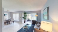 This well-appointed single-floor residence offers a thoughtfully designed layout that balances comfort and functionality. The spacious living room serves as the heart of the home, featuring light gray walls, large-format white tile flooring, and a textured ceiling. A large sliding glass door with vertical blinds opens to an outdoor area, flooding the space with natural light and providing views of lush greenery. The living area flows seamlessly into a dedicated dining space, which features a generous window overlooking the outdoors, making it an inviting area for everyday meals. The kitchen is efficiently laid out with white cabinetry, warm countertops, a full suite of appliances including a range, dishwasher, and refrigerator, and ample storage. Two well-sized bedrooms offer comfortable retreats; one features a bold accent wall and large windows, while the other is bathed in natural light from floor-to-ceiling vertical blinds. Both are finished with plush carpeting. The residence includes two full bathrooms, each with a tub-shower combination. Neutral, contemporary tones are carried throughout the entire space, creating a cohesive and calming ambiance that is both practical and visually appealing.