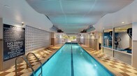 This single-level facility is purpose-built for aquatic fitness and recreation. The centerpiece is a generously proportioned indoor lap pool, finished with teal tile and flanked by a striking checkerboard-patterned surround in contrasting tones. A textured glass block wall lines one side of the pool area, diffusing natural light while maintaining privacy. The ceiling above features a sculptural, undulating design with perforated acoustic tiles and recessed lighting, contributing to both the aesthetics and acoustics of the space. At the far end of the pool, large windows and a door opening provide a view of outdoor greenery, enhancing the sense of openness.

Supporting the aquatic area are well-equipped amenity rooms. The changing room includes metal lockers with a wooden bench ledge, a curtained shower stall with grab bars, and non-slip modular flooring in blue. A beam-style scale is also present, reflecting the facility's fitness-oriented function. The restroom areas are cleanly finished with white ceramic tile flooring, granite-topped vanities, wall-mounted mirrors, and accessible fixtures including grab bars and ADA-compliant hardware throughout, ensuring the space is functional and inclusive for all users.