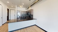Located at 612 6th St in Rapid City, South Dakota, this one-bedroom, one-bathroom unit delivers a modern industrial aesthetic across a thoughtfully designed single floor. Exposed cylindrical ductwork runs throughout the space, lending an urban loft character that is complemented by warm light wood flooring and crisp white walls.

The kitchen is a standout feature, outfitted with rich dark-toned cabinetry, a bold hexagonal tile backsplash, stainless steel appliances including a gas range and French door refrigerator, and a dark granite peninsula countertop with a sleek undermount sink. Pendant lighting above the peninsula adds a contemporary finishing touch, while the open layout keeps the kitchen connected to the main living area.

The bedroom is bright and airy, benefiting from large black-framed windows that frame expansive views of the Rapid City skyline and surrounding landscape. The same light wood flooring and clean white walls continue here, creating a calm and uncluttered retreat.

The bathroom features a full tub-shower combo with large-format tile surround, dark cabinetry with black hardware, and light wood flooring that ties the space together. The unit also includes a dedicated utility room. Bold black door frames and fixtures throughout provide strong contrast and a cohesive modern design language.