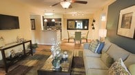 This well-appointed single-floor residence showcases an open-concept layout that effortlessly connects the living, dining, and kitchen areas into one cohesive, flowing space. Warm wood-tone flooring runs throughout, lending a sense of continuity and warmth to the interior.

The living area is anchored by a ceiling fan with a light fixture, providing both comfort and illumination. A plush sofa, decorative coffee table with an ornate tile inlay, and layered area rugs create an inviting and stylish gathering space. A large wall mirror in the adjacent dining area amplifies natural light and adds depth to the room, while a round dining table surrounded by green upholstered chairs offers an intimate setting for meals.

The kitchen is positioned at the rear of the open floor plan and features dark cabinetry, stainless steel appliances including a refrigerator and microwave, and a raised breakfast bar that serves as both a functional workspace and a casual dining option. Pendant lighting above the bar adds a modern touch.

Track lighting and strategically placed lamps ensure the space is well-lit throughout. The overall ambiance is warm, modern, and thoughtfully designed, making it a comfortable and functional home environment.