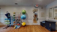 Located at 1000 Flycatcher Way in Arden, NC, this fitness center is a well-appointed, single-floor facility designed to support a wide range of workout routines. The space features warm hardwood flooring throughout, recessed lighting, and ceiling fans that contribute to a comfortable and functional exercise environment. A standout feature of the room is a dramatic floor-to-ceiling mural depicting the layered ridgelines of the Blue Ridge Mountains, adding a distinctly regional character to the space. A large wall-mounted mirror spans one side of the room, visually expanding the area and aiding in form correction during workouts. The fitness center is equipped with stationary and recumbent exercise bikes positioned near a row of windows that invite natural light and outdoor views. Organizational racks hold an assortment of resistance bands, medicine balls, stability balls in various sizes, and handheld dumbbells. A dedicated storage stand keeps yoga mats and additional accessories neatly arranged. Weight benches are available for strength training. The space is accessed through a windowed door and features clear signage, including posted fitness center guidelines. The overall ambiance is clean, organized, and welcoming, with thoughtful design elements that make it both practical and visually engaging.
