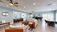 Located at 3006 Hickory Woods Dr in Roanoke, Virginia, this single-story community clubhouse offers a generously sized, open-concept layout designed for gathering and relaxation. The interior is unified by soft sky-blue walls, light wood-tone flooring, and a textured ceiling accented with recessed lighting and ceiling fans throughout.

The main living area is expansive, featuring multiple distinct seating arrangements. One lounge zone is anchored by wicker and rattan furniture grouped around a wall-mounted television, while another centers around leather and upholstered sofas paired with a wood-framed coffee table. A striking dark brick fireplace with a natural wood mantel serves as a bold architectural focal point, complemented by a mounted television above.

A coffee and beverage bar is tucked along one wall, complete with dark cabinetry, open shelving, and a mini refrigerator — ideal for casual refreshment. Adjacent to this is a bar-height counter with white barstools, offering additional seating. A dedicated workstation area with desks and computers is positioned near natural light from white-shuttered windows.

A set of elegant barn-style sliding doors leads to a separate enclosed room, adding functional versatility to the space. Large windows and glass doors throughout invite abundant natural light and views of the surrounding outdoor greenery, contributing to an open, welcoming atmosphere.