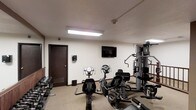 This fitness center is a dedicated single-floor exercise facility designed to accommodate a range of workout needs. The space is outfitted with multiple cardio machines, including stationary bikes, providing options for low-impact cardiovascular training. A multi-station cable and weight machine anchors the strength training area, offering versatile resistance exercise options. A rack of dumbbells lines one wall, catering to free weight workouts of varying intensity. The room is finished with neutral-toned walls and carpeted flooring, contributing to a clean and functional environment. Recessed and flush-mounted fluorescent lighting ensures the space is well-lit throughout. A wall-mounted flat-screen television is positioned for easy viewing during workouts, adding an element of entertainment. A security camera is also visible, reflecting attention to safety. The space is accessed through a dark wood-framed door, and a small waste bin is conveniently placed near the entrance. Overall, the fitness center is thoughtfully arranged to maximize usability while maintaining an organized and approachable atmosphere suited for daily exercise routines.