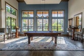 This inviting single-story clubroom in Beaumont, Texas is thoughtfully designed to serve as a premier community gathering space. Soaring ceilings with warm accent walls and elegant chandelier and pendant lighting create a welcoming ambiance throughout. The main lounge area is anchored by a full-size billiards table set atop a colorful area rug, surrounded by comfortable seating arrangements and bathed in natural light filtering through large windows. Arched entryways with stacked stone columns provide architectural distinction as they connect the various zones of the space. A cozy sitting area features a dramatic floor-to-ceiling stacked stone fireplace, accompanied by striped upholstered armchairs with accent pillows and curated shelving displaying decorative pieces. Adjacent to this lounge, large windows offer views of the outdoor area, enhancing the open, airy feel. The kitchen area is outfitted with rich dark wood cabinetry, granite countertops, a stone tile backsplash extending to the ceiling, a wall-mounted television, and appliances including a coffee station — ideal for casual entertaining. A separate dining area with a solid wood table and chairs, string lighting overhead, and direct access through French doors to the outdoors completes this well-appointed clubroom.