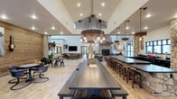 This single-floor space masterfully blends rustic charm with modern comfort, creating an inviting and functional gathering environment. The interior is anchored by soaring vaulted ceilings that rise dramatically overhead, amplifying the sense of openness and grandeur throughout. A striking barrel-style chandelier with exposed Edison bulbs serves as the centerpiece, casting warm, ambient light over a long, dark-wood communal dining table and bench seating below.

Natural stone accents are woven throughout the space, most notably in the floor-to-ceiling stone fireplace that anchors the living area, as well as in the decorative columns that frame the open floor plan. The extended bar counter, lined with wooden barstools, provides ample seating and runs the length of one side of the room, making it ideal for entertaining.

The lounge area at the far end features comfortable upholstered seating arranged around the fireplace, offering a more intimate setting within the larger open layout. Light-toned tile flooring and crisp white walls balance the richness of the dark wood and stone elements. Large windows and glass doors invite natural light deep into the space, while western-themed decor, including mounted saddles and artwork, reinforce the lodge-inspired aesthetic throughout.