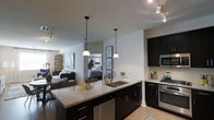 This single-floor space showcases a thoughtfully designed open-concept layout that integrates the kitchen, dining, and living areas into one cohesive environment. The kitchen serves as the focal point, featuring dark espresso cabinetry paired with granite countertops that offer ample prep and storage space. A central island with an undermount double sink provides additional workspace and acts as a natural divider between the kitchen and the adjoining living area. Pendant lighting hangs above the island, adding warmth and visual interest, while a track lighting system overhead ensures the space is well-lit throughout. Stainless steel appliances, including a gas range and microwave, complete the modern kitchen aesthetic. Light hardwood-style flooring runs throughout, creating a warm and cohesive base that complements the darker cabinet tones. The open living area beyond the kitchen is bright and airy, with natural light filtering in from windows along the far wall. A glimpse into an adjacent bedroom reveals a mirrored feature that amplifies light and adds depth. The overall ambiance is contemporary and functional, balancing clean lines with comfortable, livable design.
